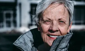 En kvinna med grått hår och korta frisyrer, runt 60 år, ler varmt. Hon bär en rutig jacka.