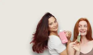 Två kvinnor med vackert hår håller upp hårprodukter. Den ena har en rosa flaska, den andra en liten grön flaska med jojobaolja för hårbotten.