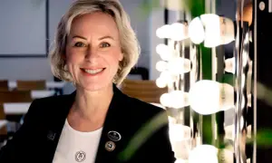 En kvinna med kort blont hår ler mot kameran. Hon bär en svart kavaj och en vit t-shirt med logotypen för Maria Åkerberg. Bakom henne syns en rad lampor.