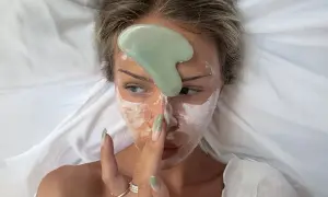 En kvinna med ansiktsmask och en grön gua sha-sten på pannan, redo för en avslappnande gua sha-massage.