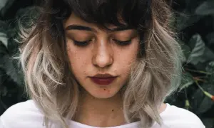 Ung kvinna med mörkbrunt hår som övergår i ljusa slingor, en look som påminner om balayage hemma. Glitter på kinden.