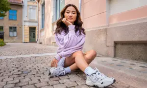 Ung kvinna med mjukt vågigt hår, en lila tröja och en "money piece" frisyr sitter på kullerstensgata.