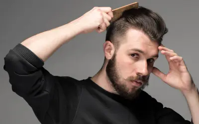 Man med skägg och mustasch stylar sitt korta hår med en kam. Frisyr med skin fade.