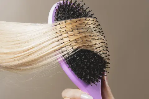 En hand med ljusa naglar håller en lila borste som används för att borsta långt, blont hår.