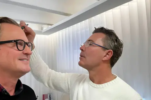 Man med glasögon får styling av sitt permanenta hår.