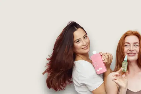 Två kvinnor med vackert hår håller upp hårprodukter. Den ena har en rosa flaska, den andra en liten grön flaska med jojobaolja för hårbotten.