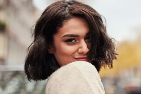 A woman with dark, wavy bob haircut and a nose ring smiles over her shoulder.