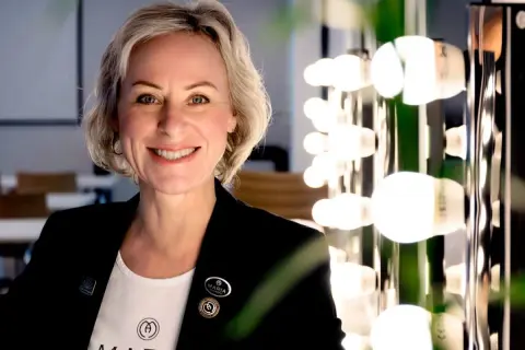 En kvinna med kort blont hår ler mot kameran. Hon bär en svart kavaj och en vit t-shirt med logotypen för Maria Åkerberg. Bakom henne syns en rad lampor.