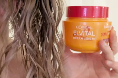 En hand håller en burk med L'Oréal Elvital Dream Length hårinpackning bredvid blont, vått hår.