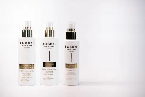 Tre flaskor Bobby's Hair Care i vitt med gulddetaljer. En leave-in conditioner för värmeskydd, en saltspray för beach look och en scalp elixir med rosmarindoft.
