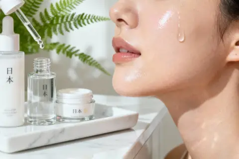 En droppe serum faller mot en glänsande hud, omgiven av japanska skönhetsprodukter. J beauty för en strålande hy.