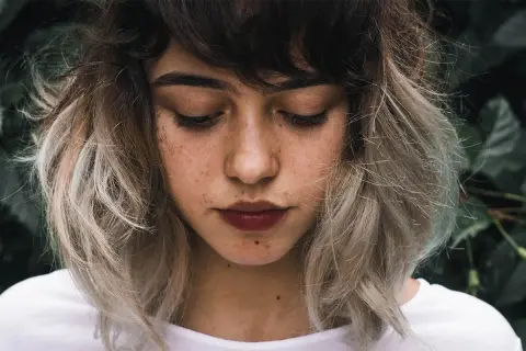 Ung kvinna med mörkbrunt hår som övergår i ljusa slingor, en look som påminner om balayage hemma. Glitter på kinden.