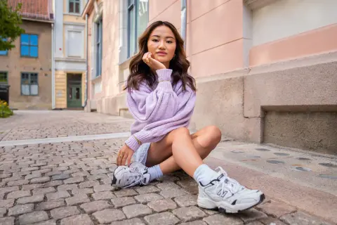 Ung kvinna med mjukt vågigt hår, en lila tröja och en "money piece" frisyr sitter på kullerstensgata.