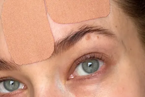 Närbild på ögon och panna med hudfärgad face taping. Ögonen är gröna och har långa fransar.