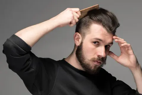 Man med skägg och mustasch stylar sitt korta hår med en kam. Frisyr med skin fade.