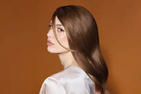 En kvinna med glansigt brunt hår och en subtil nyansering hemma, blickar bort med en mjuk makeup.