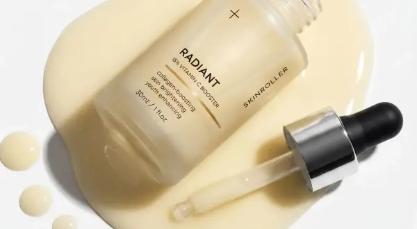 En flaska med "RADIANT 15% VITAMIN C BOOSTER" serum ligger på en gulaktig vätska. Pipetten har en droppe pha syra redo att appliceras.