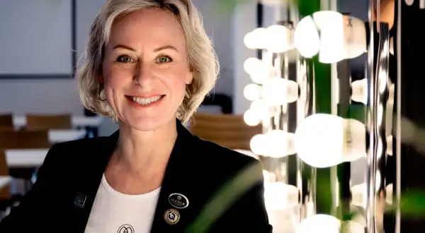 En kvinna med kort blont hår ler mot kameran. Hon bär en svart kavaj och en vit t-shirt med logotypen för Maria Åkerberg. Bakom henne syns en rad lampor.