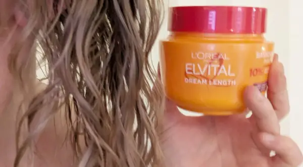 En hand håller en burk med L'Oréal Elvital Dream Length hårinpackning bredvid blont, vått hår.
