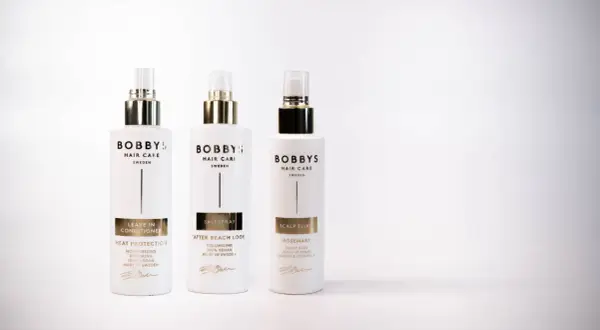 Tre flaskor Bobby's Hair Care i vitt med gulddetaljer. En leave-in conditioner för värmeskydd, en saltspray för beach look och en scalp elixir med rosmarindoft.