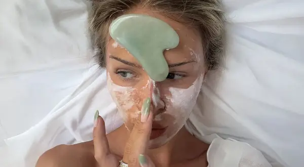 En kvinna med ansiktsmask och en grön gua sha-sten på pannan, redo för en avslappnande gua sha-massage.