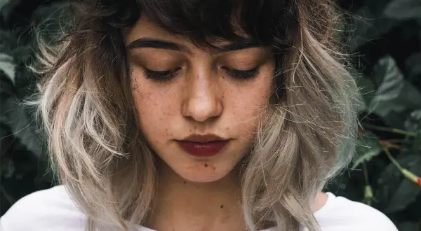 Ung kvinna med mörkbrunt hår som övergår i ljusa slingor, en look som påminner om balayage hemma. Glitter på kinden.