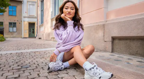Ung kvinna med mjukt vågigt hår, en lila tröja och en "money piece" frisyr sitter på kullerstensgata.