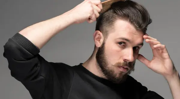 Man med skägg och mustasch stylar sitt korta hår med en kam. Frisyr med skin fade.
