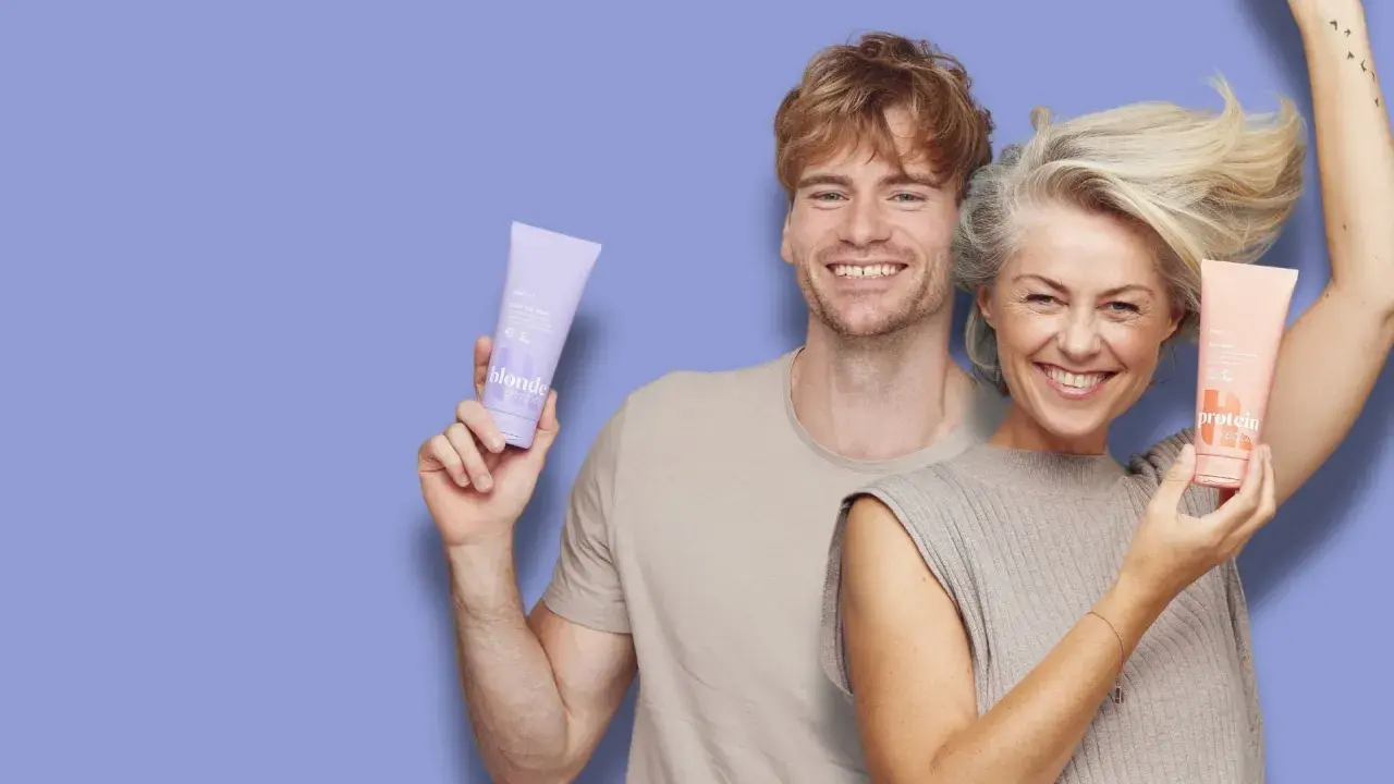 Glad man och kvinna ler och håller upp hårinpackningar. Hon har protein repair, han har blonde.