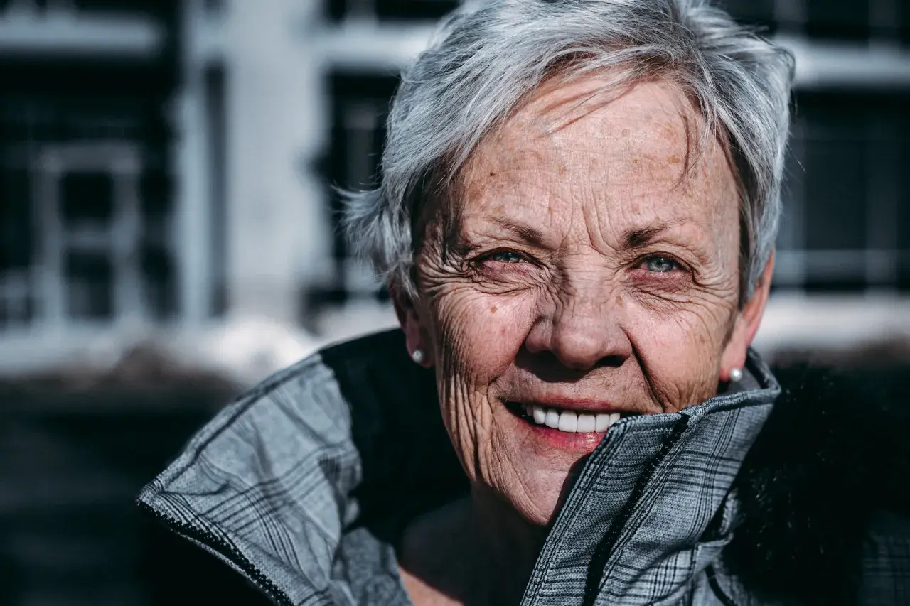 En kvinna med grått hår och korta frisyrer, runt 60 år, ler varmt. Hon bär en rutig jacka.
