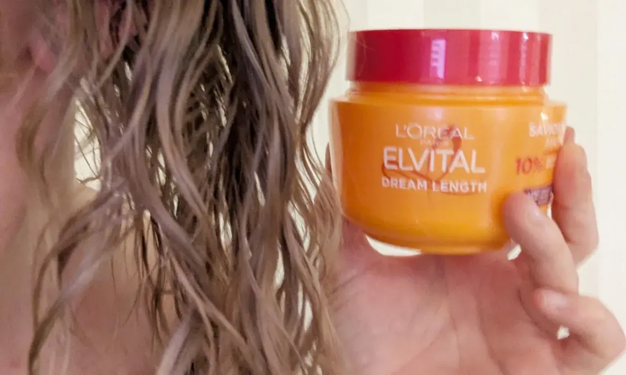 En hand håller en burk med L'Oréal Elvital Dream Length hårinpackning bredvid blont, vått hår.