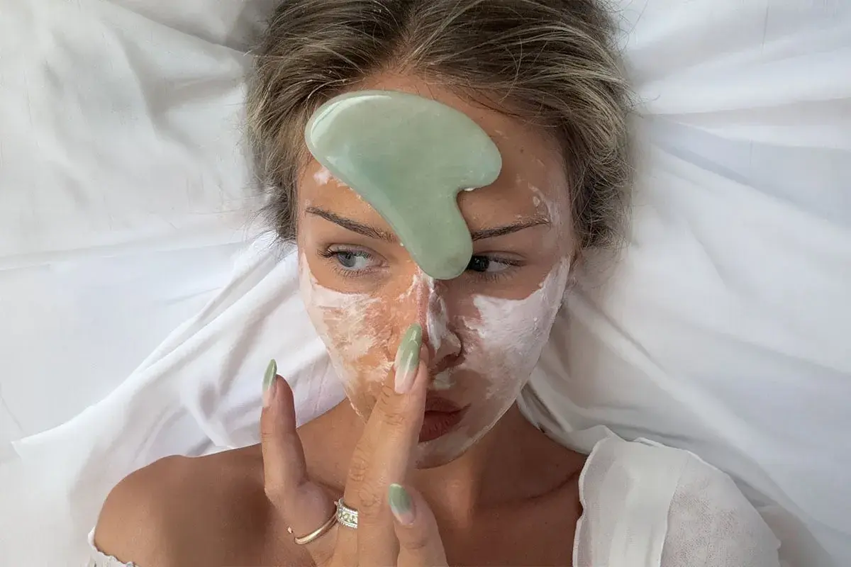 En kvinna med ansiktsmask och en grön gua sha-sten på pannan, redo för en avslappnande gua sha-massage.