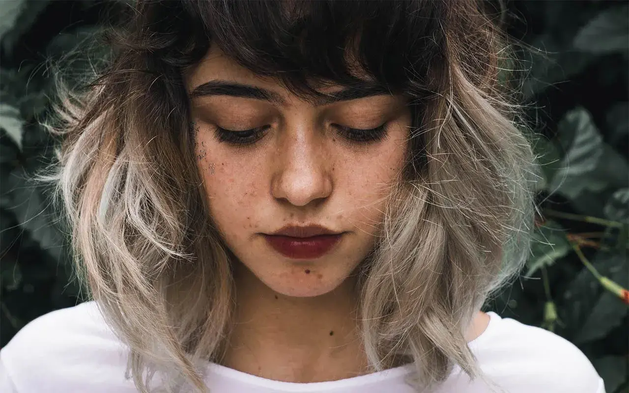 Ung kvinna med mörkbrunt hår som övergår i ljusa slingor, en look som påminner om balayage hemma. Glitter på kinden.