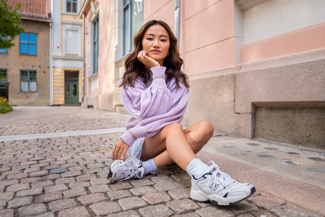 Ung kvinna med mjukt vågigt hår, en lila tröja och en "money piece" frisyr sitter på kullerstensgata.