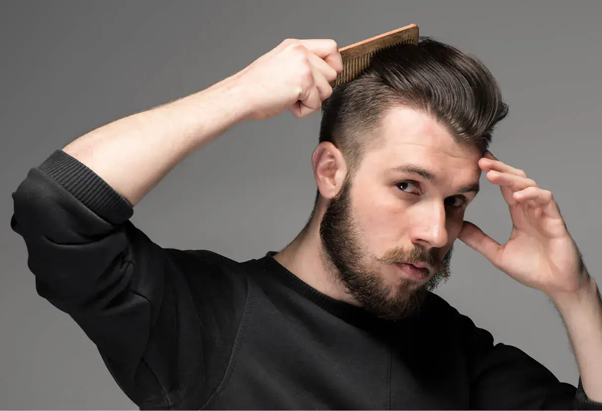 Man med skägg och mustasch stylar sitt korta hår med en kam. Frisyr med skin fade.