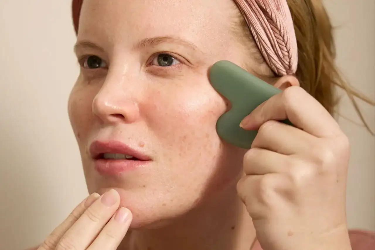 Kvinna med rosa pannband använder en grön gua sha för ansiktsmassage.