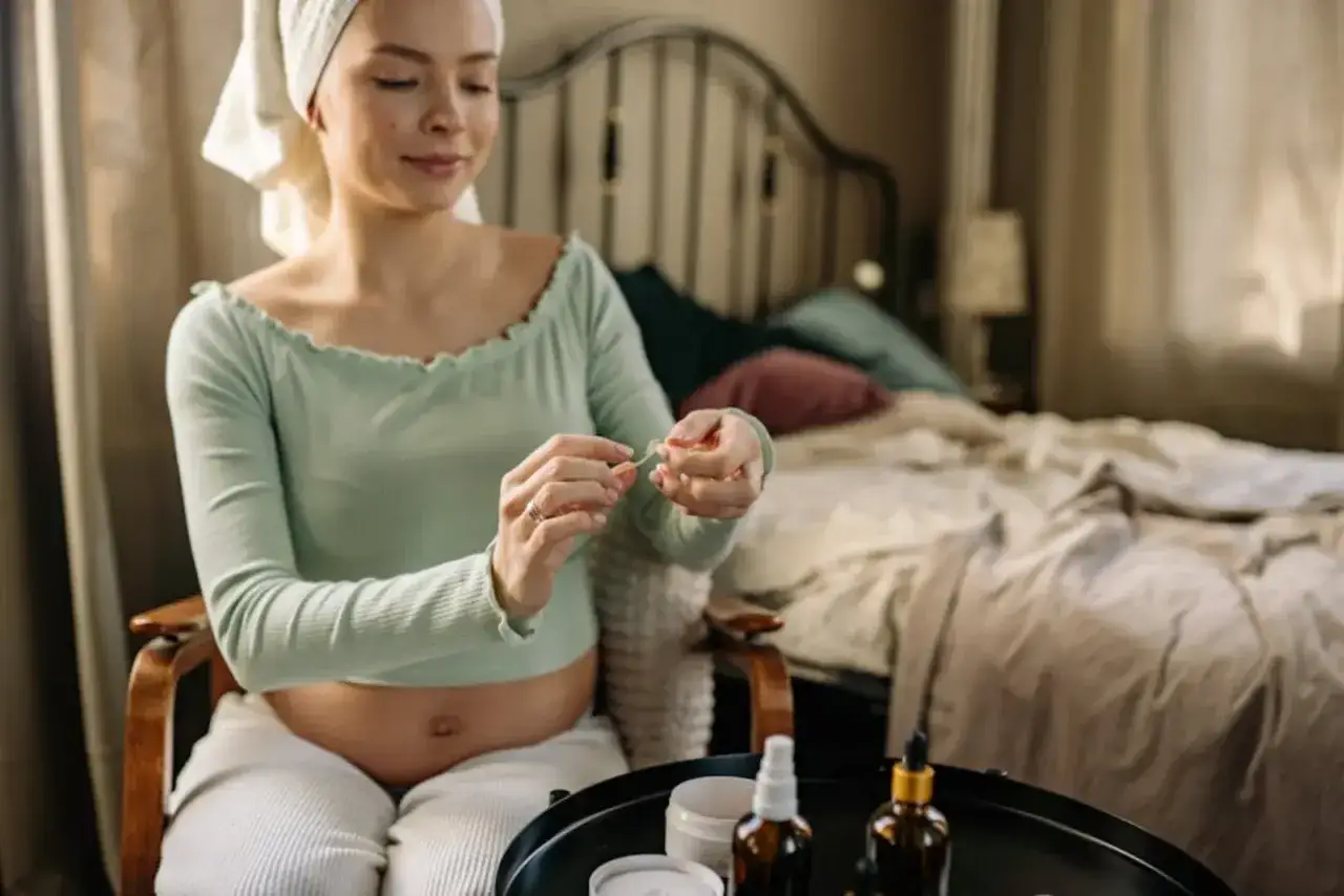 Gravid kvinna med handduk på huvudet applicerar en ansiktsmask. Hon sitter i en stol med en säng i bakgrunden.