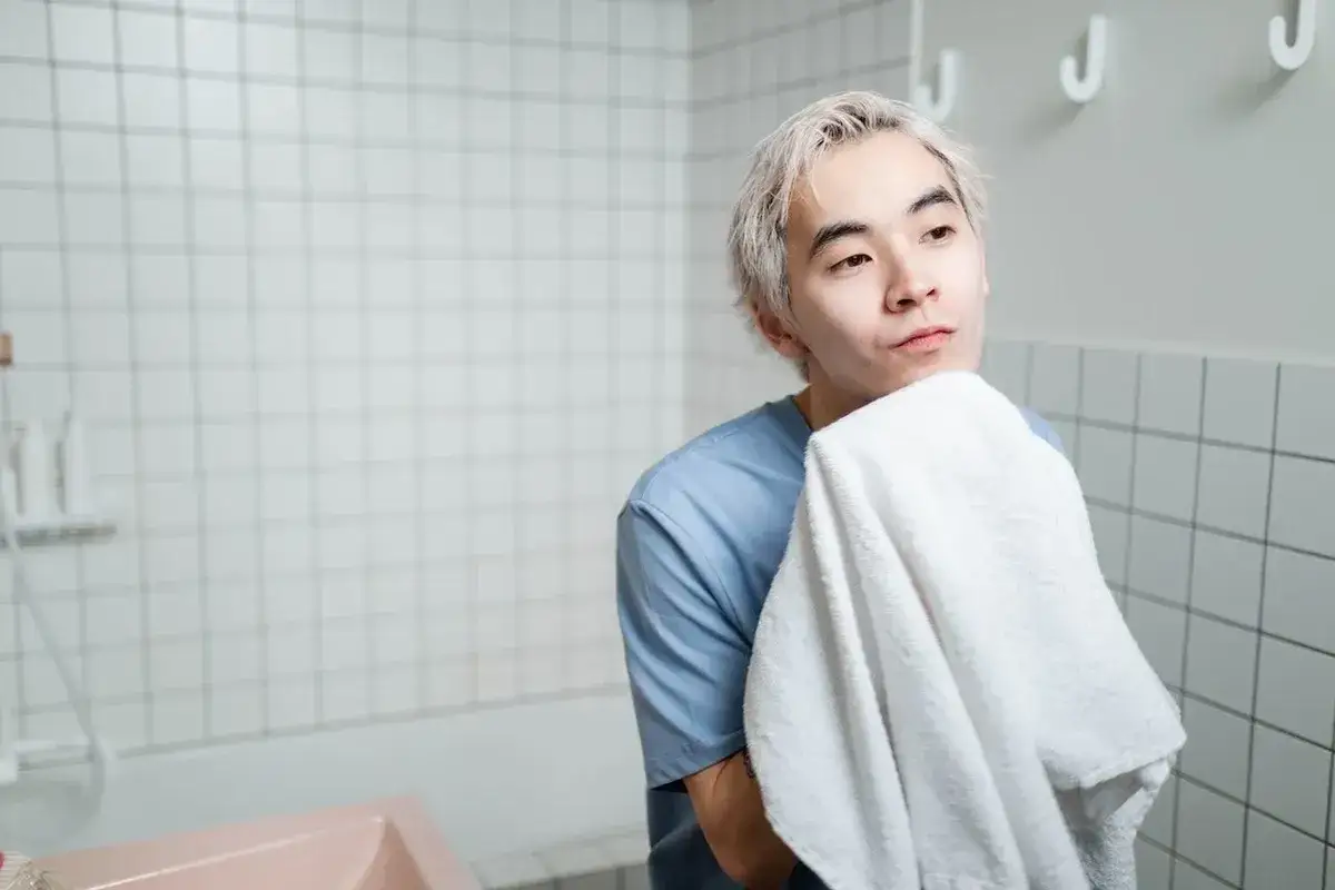En ung man med blont hår torkar sitt ansikte med en vit handduk. Han har en blå tröja på sig och står i ett badrum med kaklade väggar. Hans hud ser lite fet ut.