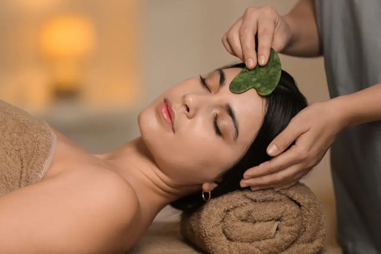 En kvinna f&aring;r en avslappnande gua sha massage i ansiktet. En gr&ouml;n sten glider &ouml;ver hennes panna.