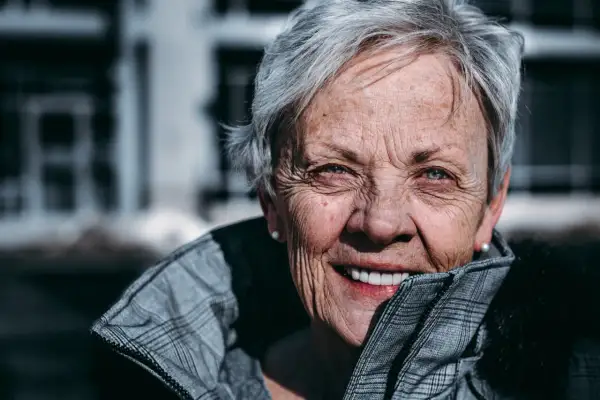 En kvinna med grått hår och korta frisyrer, runt 60 år, ler varmt. Hon bär en rutig jacka.