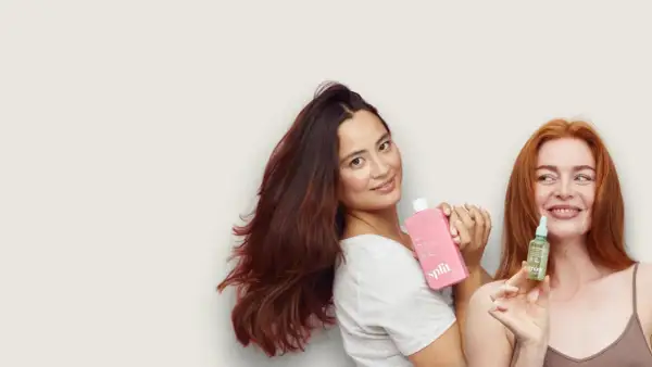 Två kvinnor med vackert hår håller upp hårprodukter. Den ena har en rosa flaska, den andra en liten grön flaska med jojobaolja för hårbotten.
