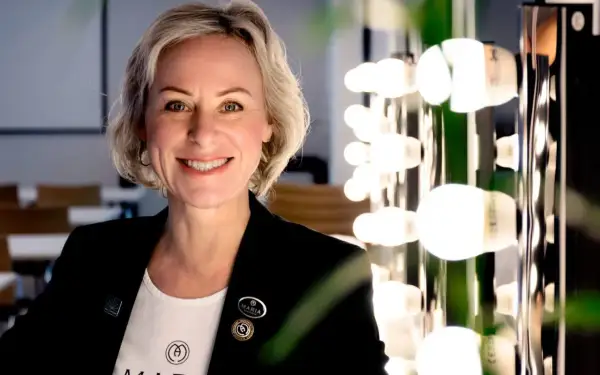 En kvinna med kort blont hår ler mot kameran. Hon bär en svart kavaj och en vit t-shirt med logotypen för Maria Åkerberg. Bakom henne syns en rad lampor.