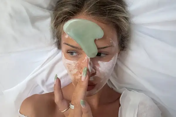 En kvinna med ansiktsmask och en grön gua sha-sten på pannan, redo för en avslappnande gua sha-massage.