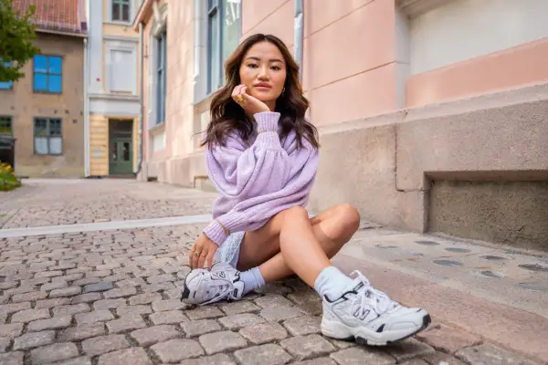 Ung kvinna med mjukt vågigt hår, en lila tröja och en "money piece" frisyr sitter på kullerstensgata.